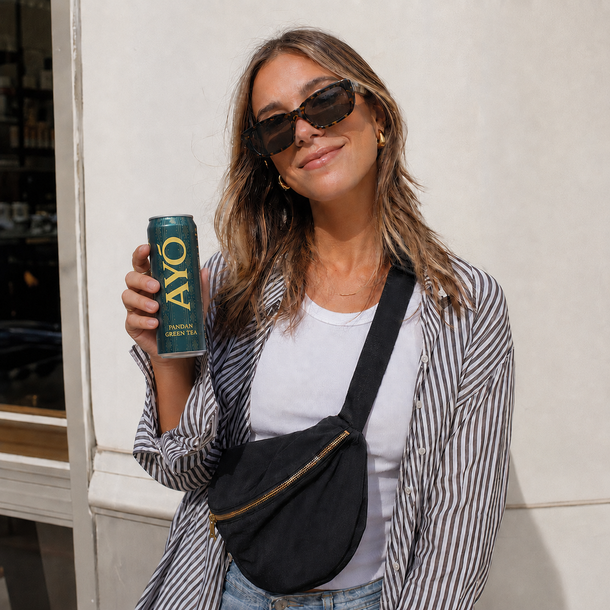 Woman in sunglasses holding a cold AYÓ Pandan Green Tea can on a city sidewalk